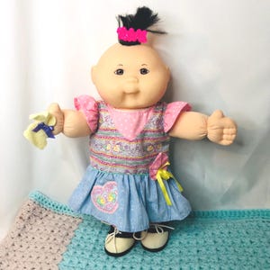 May include: A vintage Cabbage Patch Kid doll with a bald head and a tuft of black hair held by a pink bow. The doll wears a pink and blue dress with a heart-shaped patch and black and white shoes. It holds a yellow cloth with a blue bow.