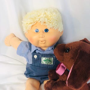 Puede incluir: Una muñeca Cabbage Patch Kids vintage con cabello rubio de hilo, ojos azules y mejillas rosadas, vestida con una camisa azul a cuadros y un peto vaquero. Un perro de peluche marrón con un collar rosa está al lado de la muñeca.