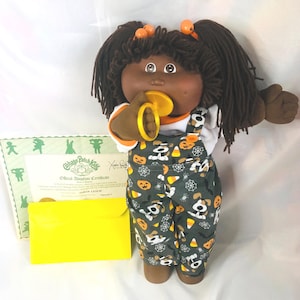 Op de afbeelding: Een Cabbage Patch Kids pop met donkerbruin wollen haar in staartjes, gekleed in een wit shirt en een donkergrijze tuinbroek met een Halloween-print van pompoenen, maissnoepjes en spinnenwebben. De pop houdt een gele fopspeen vast en wordt geleverd met een adoptiecertificaat en een gele envelop.