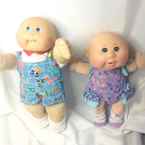 May include: Two vintage Cabbage Patch Kids dolls. One doll wears a blue and white patterned outfit with a red collar. The other doll wears a purple floral dress with blue trim. Both dolls have bald heads, blue or green eyes, and are in a standing position.