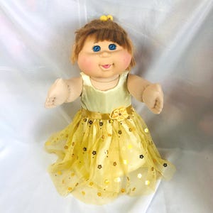 Op de afbeelding: Een pop met bruin garen haar, blauwe ogen en roze wangen draagt een gele mouwloze jurk. De jurk heeft een satijnen lijfje, een tailleband met een stoffen bloem en een tule rok versierd met kleine gouden en bruine bloemvormige lovertjes.