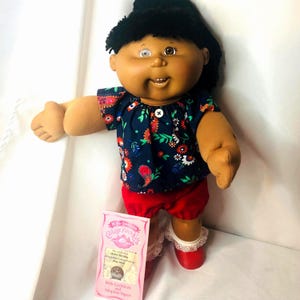 May include: A Cabbage Patch Kids doll with dark hair, brown skin, and one light-colored eye. The doll wears a navy blue top with a colorful floral and paisley pattern, a single white button, red shorts, and red shoes with white lace trim.