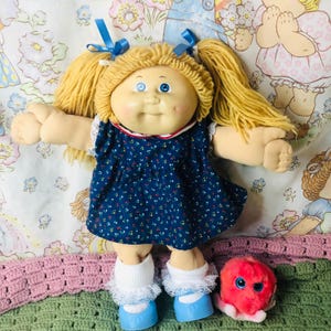 May include: A vintage Cabbage Patch Kid doll with blonde yarn pigtails, blue eyes, and a rosy complexion. The doll wears a blue floral dress, white socks with lace trim, and blue shoes. A small pink plush toy with blue eyes sits nearby.