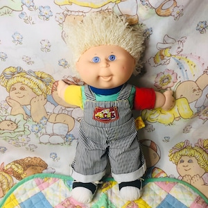 Könnte beinhalten: Eine Vintage-Cabbage-Patch-Kids-Puppe mit blondem, lockigem Haar und blauen Augen. Die Puppe trägt ein blaues, grünes, rotes und gelbes Hemd unter einer gestreiften Latzhose. Die Latzhose hat einen Aufnäher mit der Aufschrift "Lucas". Die Puppe steht auf einem bunten Quilt.