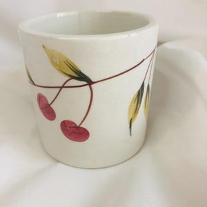 May include: A white ceramic cup with a hand-painted cherry design. The cup features two red cherries, brown stems, and yellow and green leaves. The cup has a cylindrical shape and a slightly cracked surface.