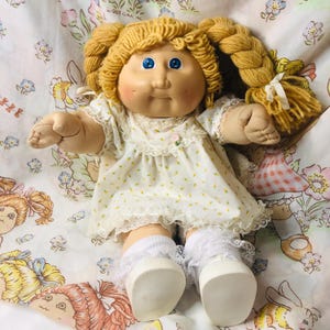 May include: A vintage Cabbage Patch Kid doll with blonde yarn pigtails and blue eyes. The doll wears a white dress with yellow floral print and lace trim, white socks, and white shoes. The doll is on a floral patterned fabric.