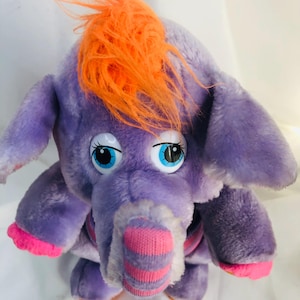 Op de afbeelding: Een paars pluche olifantenspeelgoed met oranje haar en grote blauwe ogen. De olifant heeft een roze en wit gestreepte slurf, roze voeten en een roze kraag. Dit knuffeldier is een vintage speelgoed.