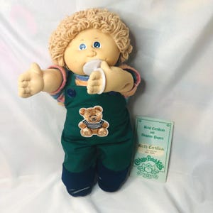 May include: A Cabbage Patch Kids doll with blonde yarn hair, blue eyes, and a white pacifier. The doll wears dark green overalls with a brown teddy bear patch and a striped shirt. Beside the doll is a light green birth certificate and adoption papers.