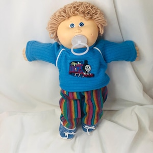 Peut inclure: Poupée vintage avec des cheveux blonds en fil, des yeux bleus et une sucette. La poupée porte un pull bleu avec un écusson Thomas le petit train, un pantalon rayé et des chaussures bleues. Les bras de la poupée sont tendus.