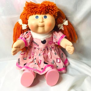 May include: A doll with bright orange yarn hair styled in pigtails with white bows, blue eyes, and rosy cheeks. The doll wears a pink dress featuring a repeating pattern of black and white Barbie-themed graphics and the text Barbie Girl.
