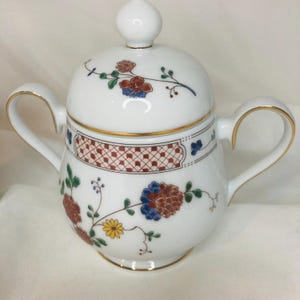 Może przedstawiać: Biała porcelanowa cukiernica z pokrywką, zdobiona kwiatowymi wzorami w kolorze czerwonym, niebieskim i żółtym. Cukiernica ma dwa uchwyty i złote obramowanie. Pokrywka zwieńczona jest małym, białym guzikiem.