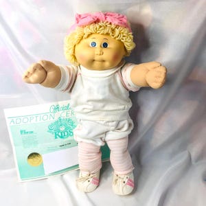 Op de afbeelding: Een Cabbage Patch Kids pop met blond garen haar, een roze hoofdband en blauwe ogen. De pop draagt een wit shirt met korte mouwen, een witte korte broek, een roze-witte gestreepte legging en witte sneakers met roze accenten.