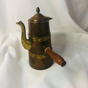 Puede incluir: Cafetera antigua de cobre con detalles de latón y un asa de madera. La cafetera tiene un pico curvo, una tapa con un adorno decorativo y bandas de latón alrededor del cuerpo. Un artículo de cocina clásico.