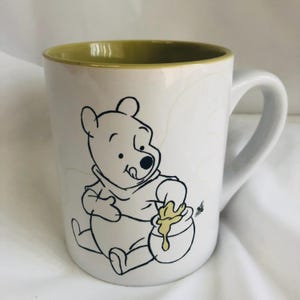 Puede incluir: Taza de cerámica blanca con una ilustración en contorno negro de Winnie the Pooh alcanzando un tarro de miel. La taza tiene un interior verde y un asa blanca. También se representa una pequeña abeja.