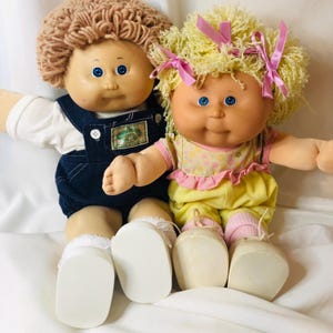 Könnte beinhalten: Zwei Puppen mit weichen Körpern und Vinylköpfen. Eine Puppe hat braunes Wollhaar, blaue Augen, ein weißes Hemd und eine dunkelblaue Jeanslatzhose. Die andere Puppe hat gelbes Wollhaar mit rosa Schleifen, blaue Augen und ein geblümtes Oberteil.