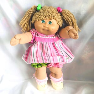 Può includere: Una bambola con capelli in lana marrone chiaro acconciati in codini con perline verdi e rosa. La bambola ha occhi verdi e indossa un vestito a righe rosa e bianche con colletto bianco, mutandoni verdi, calzini rosa a balze e scarpe color crema.