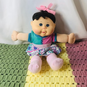 Könnte beinhalten: Eine Vintage-Puppe mit schwarzen Haaren, blauen Augen und rosigen Wangen. Die Puppe trägt ein türkis-rosa Oberteil, einen gemusterten Rock und rosa Socken. Eine rosa Schleife ziert den Kopf der Puppe. Die Puppe sitzt auf einer bunten Häkeldecke.