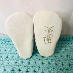 Può includere: Un paio di scarpine bianche, suole rivolte verso l'alto, con superficie strutturata. La scarpa destra ha un testo dorato che recita "Xian". Le scarpe poggiano su una coperta all'uncinetto azzurra.