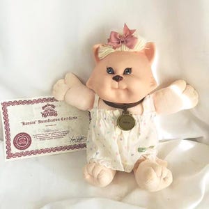 Peut inclure: Poupée chat Koosas vintage avec un nœud rose, vêtue d'un bloomer blanc à motifs de cœurs. La poupée a un collier marron avec une étiquette dorée et tend les bras. Un certificat d'identification est visible.
