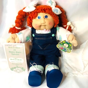 May include: A Cabbage Patch Kids doll with bright orange yarn hair in pigtails, blue eyes, and rosy cheeks. The doll wears a white long-sleeved shirt with a colorful pattern, dark blue denim overalls, and blue shoes. Includes a birth certificate.