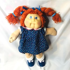 May include: A doll with orange yarn hair styled in two braided pigtails tied with blue ribbons. The doll has a round face with rosy cheeks, blue eyes, and a small mouth. It wears a dark blue dress with a tiny floral pattern, a white collar, and white socks with dark blue shoes.