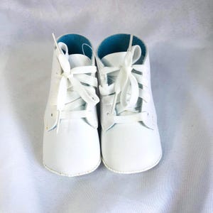 May include: A pair of white ankle boots featuring a smooth exterior, white laces tied in bows, and a bright turquoise interior lining. The boots have a rounded toe design and are constructed with visible stitching along the sides and base.