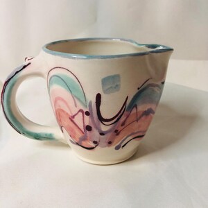 May include: Cream-colored ceramic pitcher with a rounded handle and a spout. The pitcher is decorated with abstract watercolor designs in shades of pink, teal, purple, and black. The rim is outlined in light blue.