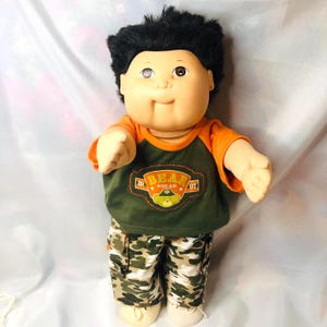 May include: A doll with a round face, tan skin, and short black hair. It wears an orange and olive green raglan shirt with a graphic patch that reads BEAR SQUAD 1997, featuring a bear icon. The doll also wears camouflage cargo pants and white shoes.