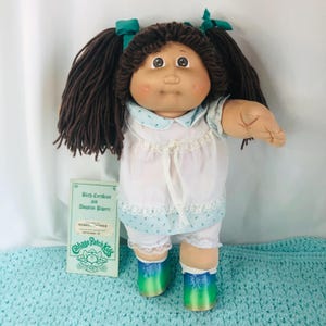 Pode incluir: Uma boneca Cabbage Patch Kids vintage com rabos de cavalo de fio castanho, vestindo um vestido branco com detalhes florais azuis e bloomers combinando. A boneca tem sapatos azuis e verdes e uma certidão de nascimento. É um item de colecionador.