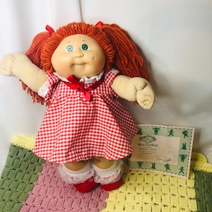 Może przedstawiać: Zabytkowa lalka Cabbage Patch Kids z rudymi włosami w warkoczach. Lalka ma na sobie czerwoną i białą sukienkę w kratkę, czerwone buty i białe skarpetki z koronkowym wykończeniem. Dołączony jest akt urodzenia.