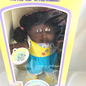 Op de afbeelding: Een Cabbage Patch Kids Toddler pop met een donkere huid en zwart garen haar in staartjes, gekleed in een wit shirt met een kleurrijk hartjespatroon, een geel vestje met twee knopen en een blauwe korte broek, verpakt in een doos met de tekst Toddler Kids.
