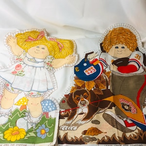 May include: Two Cabbage Patch Kids fabric panels. One panel features a girl in a floral dress, the other a boy in a football uniform holding a football. A beagle dog is also featured. The panels are ready for stuffing.