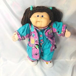 Pode incluir: Uma boneca com cabelo de lã castanho em maria-chiquinhas com elásticos turquesa, olhos azuis e rosto de vinil macio. A boneca usa um casaco com padrão de grelha turquesa e roxo sobre um macacão turquesa a condizer com botões cor-de-rosa.