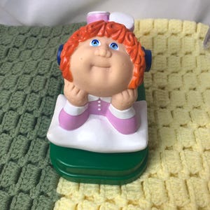 May include: A vintage Cabbage Patch Kids figurine. The doll has orange hair, blue eyes, and is wearing a pink and white outfit. The figurine is on a green base.