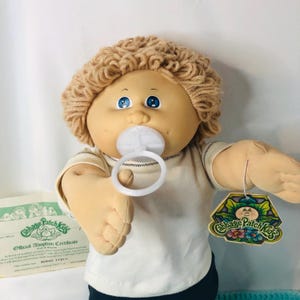 Può includere: Bambola Cabbage Patch Kids con capelli biondi di filo, occhi blu e un ciuccio. La bambola indossa una maglietta bianca e ha un'etichetta attaccata. Un certificato di adozione è visibile sullo sfondo.