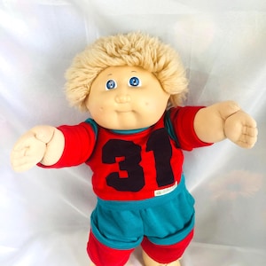 Puede incluir: Una muñeca de peluche con cabello de lana rubia y ojos azules, que viste una camiseta roja con el número 31 en negro en la parte delantera, combinada con pantalones cortos de color verde azulado con puños rojos. Tiene un cuerpo suave y brazos extendidos.