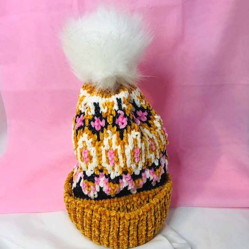 Peut inclure: Un bonnet tricot&eacute; fait main avec un pompon en fausse fourrure blanche. Le bonnet pr&eacute;sente un motif de jaune moutarde, blanc, noir et rose. Le bord est de couleur jaune moutarde uni. Le bonnet est con&ccedil;u pour le froid.