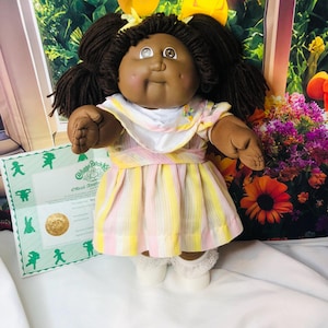 Könnte beinhalten: Eine Cabbage Patch Kids Puppe mit dunkelbraunem Wollhaar in Zöpfen, brauner Haut und braunen Augen. Die Puppe trägt ein weiß-gelb-rosa gestreiftes Kleid mit weißem Kragen und weißen Socken mit Spitzenbesatz sowie ein offizielles Adoptionszertifikat.