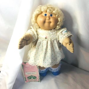 Peut inclure: Une poupée Cabbage Patch Kids aux cheveux blonds bouclés et aux yeux bleus, vêtue d'une robe blanche à petits motifs floraux bleus avec une bordure en dentelle. La poupée a un corps en tissu souple et est accompagnée d'un certificat de naissance rose.