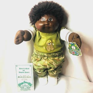 Pode incluir: Uma boneca Cabbage Patch Kids de pele escura e cabelo castanho escuro, vestindo uma camisa verde com um gráfico de urso e calças camufladas. A boneca tem uma camisa branca de manga comprida, sapatos brancos e uma etiqueta. Certificado.