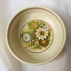 May include: Cream-colored ceramic bowl with a floral design. The center of the bowl features a circular pattern of white, yellow, and purple flowers with green leaves. A brown line borders the floral design and the rim of the bowl.
