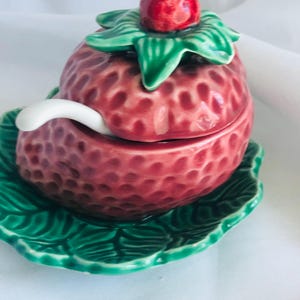 Puede incluir: Un recipiente de cerámica con forma de fresa con tapa y una cuchara pequeña, sobre un plato verde con forma de hoja. El recipiente es rojo vibrante con una superficie texturizada, y la tapa está rematada con una fresa roja y hojas verdes.