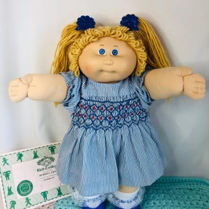 Könnte beinhalten: Eine Vintage Cabbage Patch Kids Puppe mit blonden Wollhaaren in Zöpfen, blauen Augen und einem weichen, runden Gesicht. Die Puppe trägt ein blau-weiß gestreiftes Kleid mit roten Akzenten und blaue Schuhe. Eine Geburtsurkunde ist sichtbar.