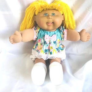May include: A doll with bright yellow yarn hair, green eyes, and a soft fabric face. It wears a teal and white patterned top featuring colorful flowers and a small pink bow at the center, paired with white lace-trimmed bloomers and white shoes.