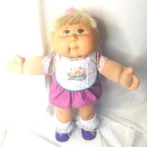 May include: A vintage Cabbage Patch Kids doll with blonde hair and blue eyes. The doll wears a pink and white floral print top, a purple skirt, and white socks with lace trim. The doll's bib has a graphic of three cartoon characters.