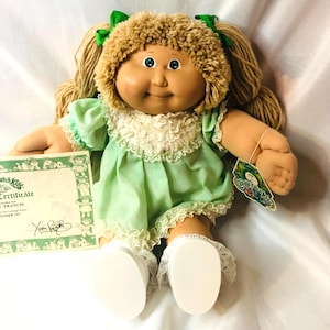 May include: A Cabbage Patch Kids doll with tan skin, green eyes, and light brown yarn hair styled in pigtails with green ribbons. The doll wears a light green dress with a white lace collar and white shoes, accompanied by a paper certificate.