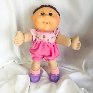 Könnte beinhalten: Eine Vintage-Puppe mit rundem Gesicht, braunen Haaren und großen Augen. Die Puppe trägt ein rosa Hemd mit Knöpfen und Nähmaschinenmotiven, rosa Shorts, rosa Socken und lila Schuhe. Die Arme der Puppe sind ausgestreckt.