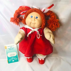 Puede incluir: Una muñeca Cabbage Patch Kids con cabello de lana naranja peinado en dos trenzas con cintas rojas y blancas. La muñeca tiene ojos azules, un vestido rojo con cuello y lazo blanco, y zapatos rojos. A su lado hay una tarjeta de certificado de nacimiento.