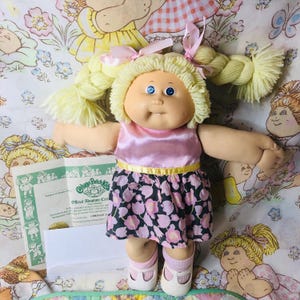 Puede incluir: Muñeca Cabbage Patch Kids con trenzas rubias de hilo, vestida con una blusa rosa satinada, una falda negra con estampado floral rosa y zapatos blancos. La muñeca tiene ojos azules y sostiene un certificado de adopción.