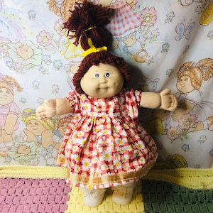 Könnte beinhalten: Eine Vintage Cabbage Patch Kid Puppe mit braunem Garnhaar, das mit einem gelben Band zusammengebunden ist. Die Puppe trägt ein rot-weiß kariertes Kleid mit Gänseblümchen-Akzenten und weißen Schuhen. Die Puppe hat blaue Augen und ein weiches, abgerundetes Gesicht.